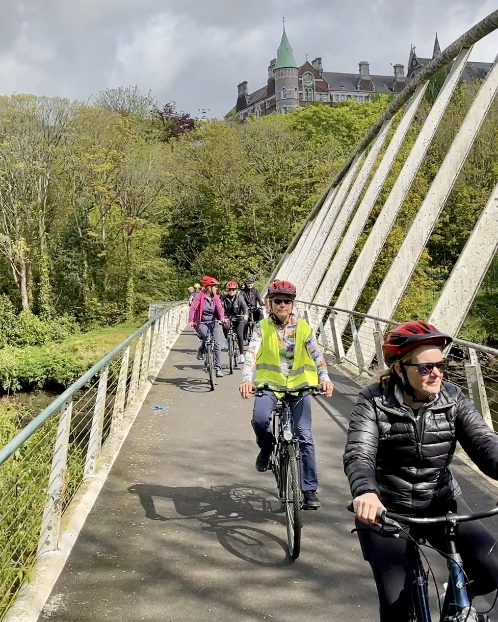 A guided group cycling across a footbridge in Cork with trees and a historic building behind them