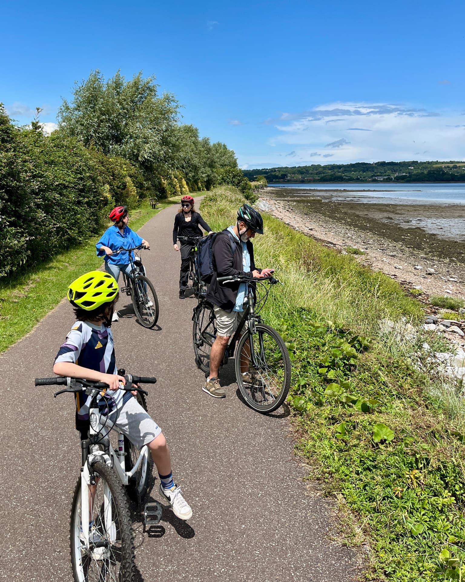 Four cyclists stopped on the waterside Greenway path beside Cork Harbour, with the tidal shoreline and green banks alongside