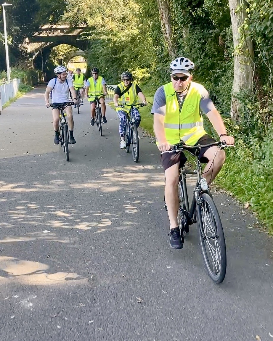 Guided cycling group in high-vis vests riding along the tree-lined former railway Greenway, with old railway arches behind them