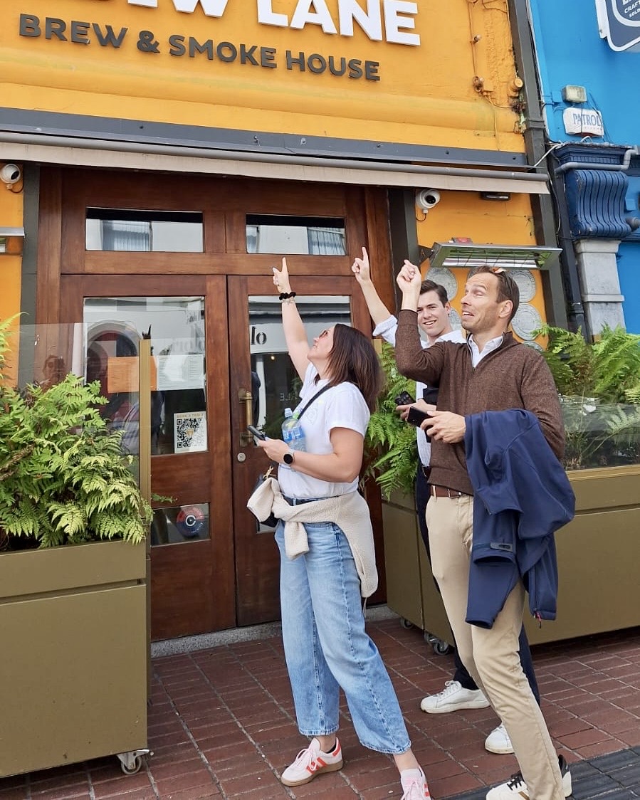 Three people pointing upwards at a building while taking part in a Cork City scavenger hunt outside a city-centre premises